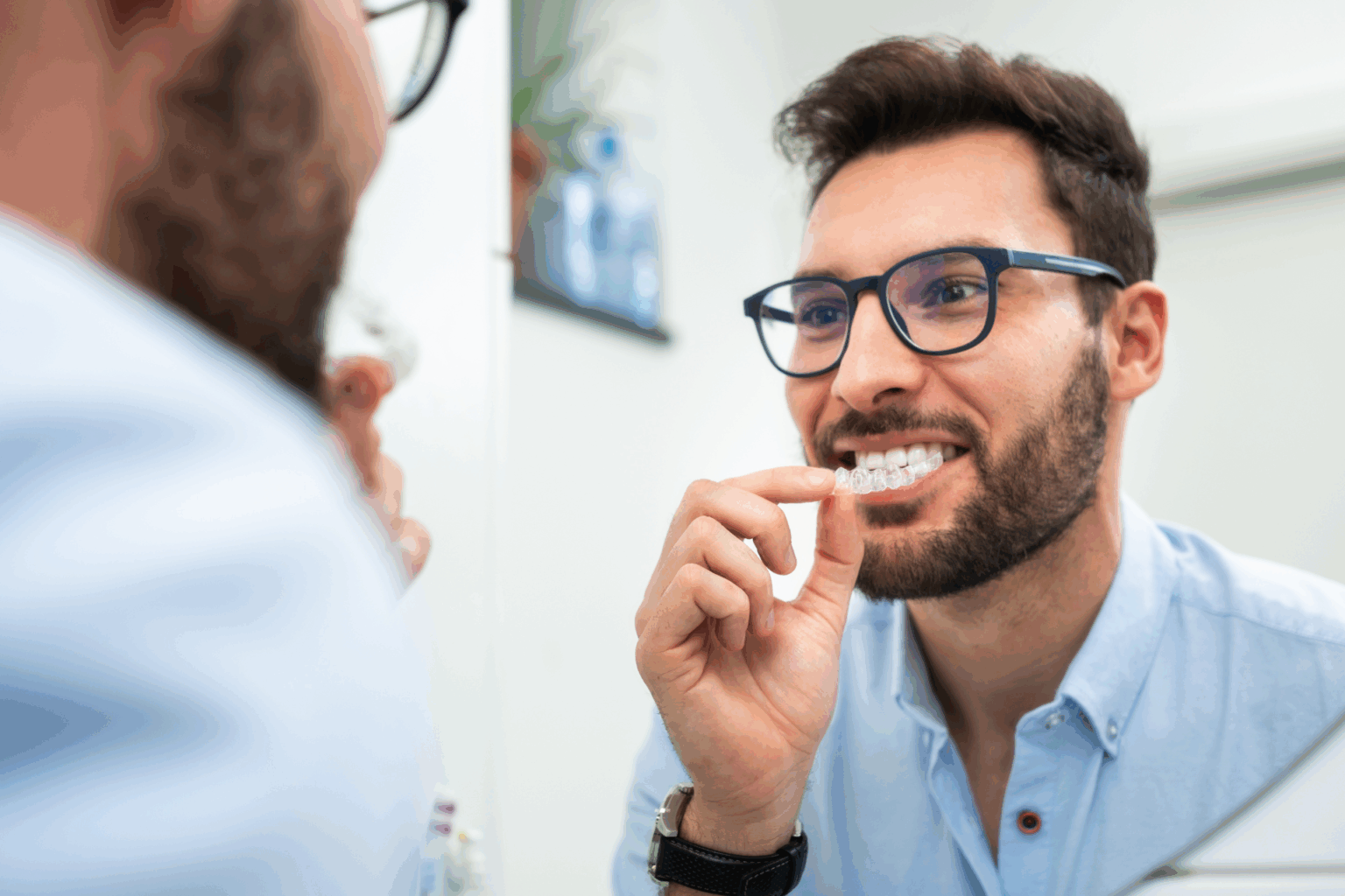 A man with glasses smiling in a mirror while holding a clear aligner